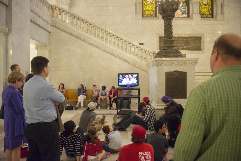 An overflow of people attending a public hearing for a proposed $15 minimum wage ordinance sit on the ground floor of Minneapolis City Hall watching the hearing take place live.  